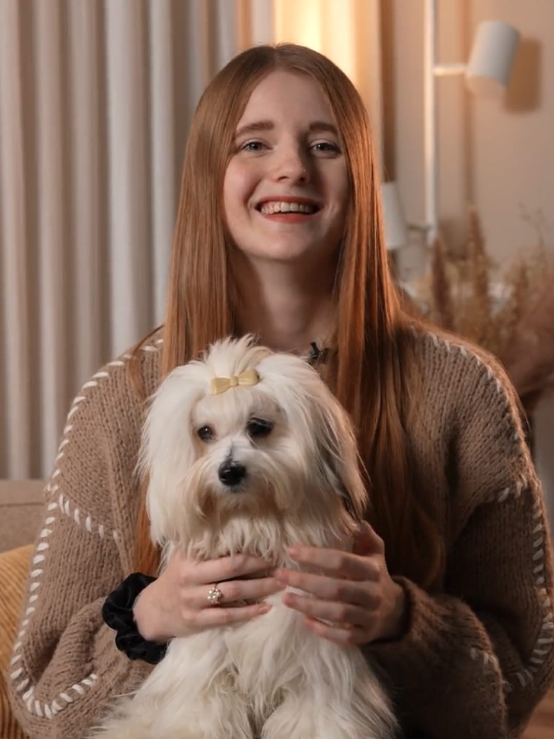 Smilende ung kvinde med langt rødt hår holder lille hvid hund med gul sløjfe