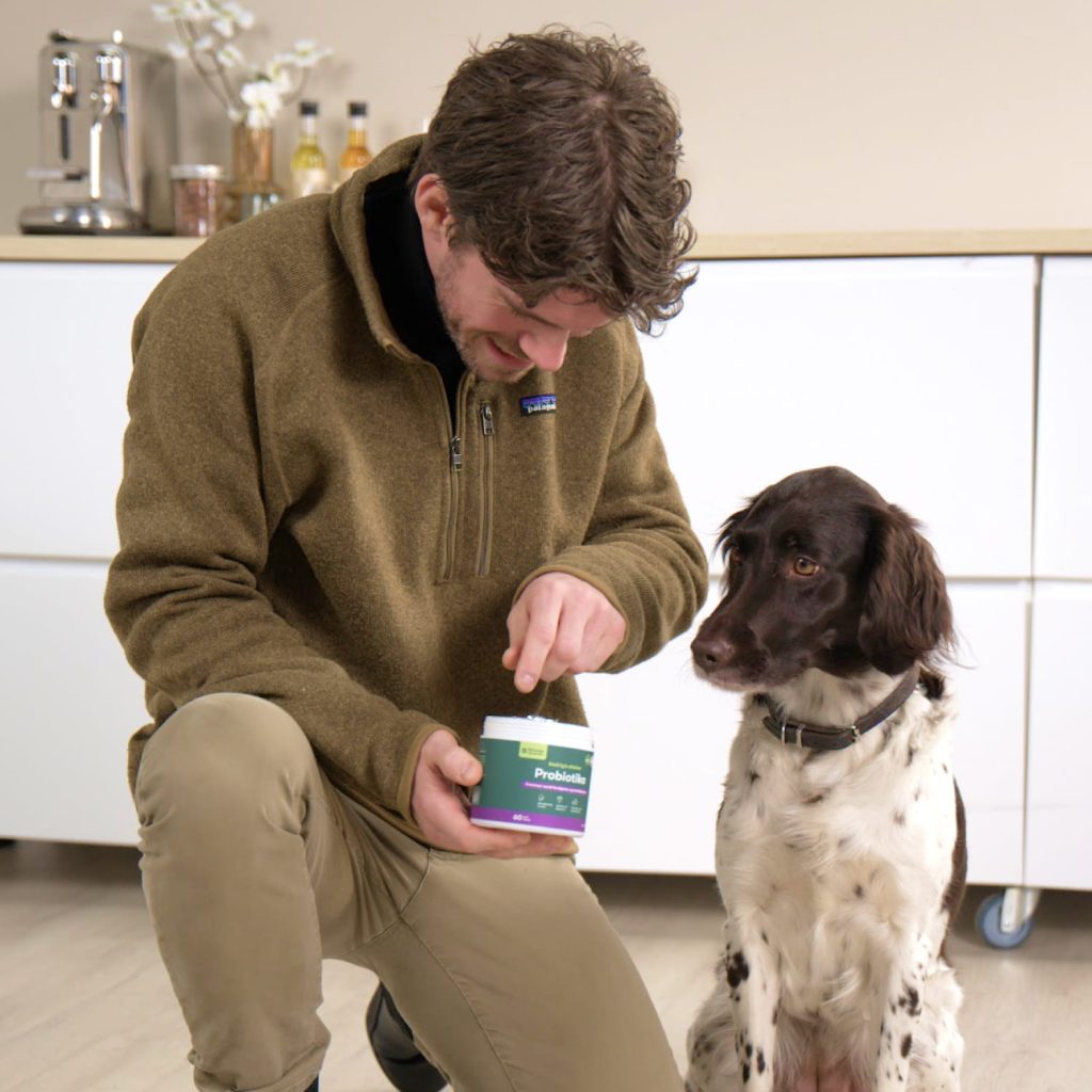 Mand i brun fleecejakke holder probiotikabøtte, mens en brun-hvid hund ser interesseret til i køkkenet